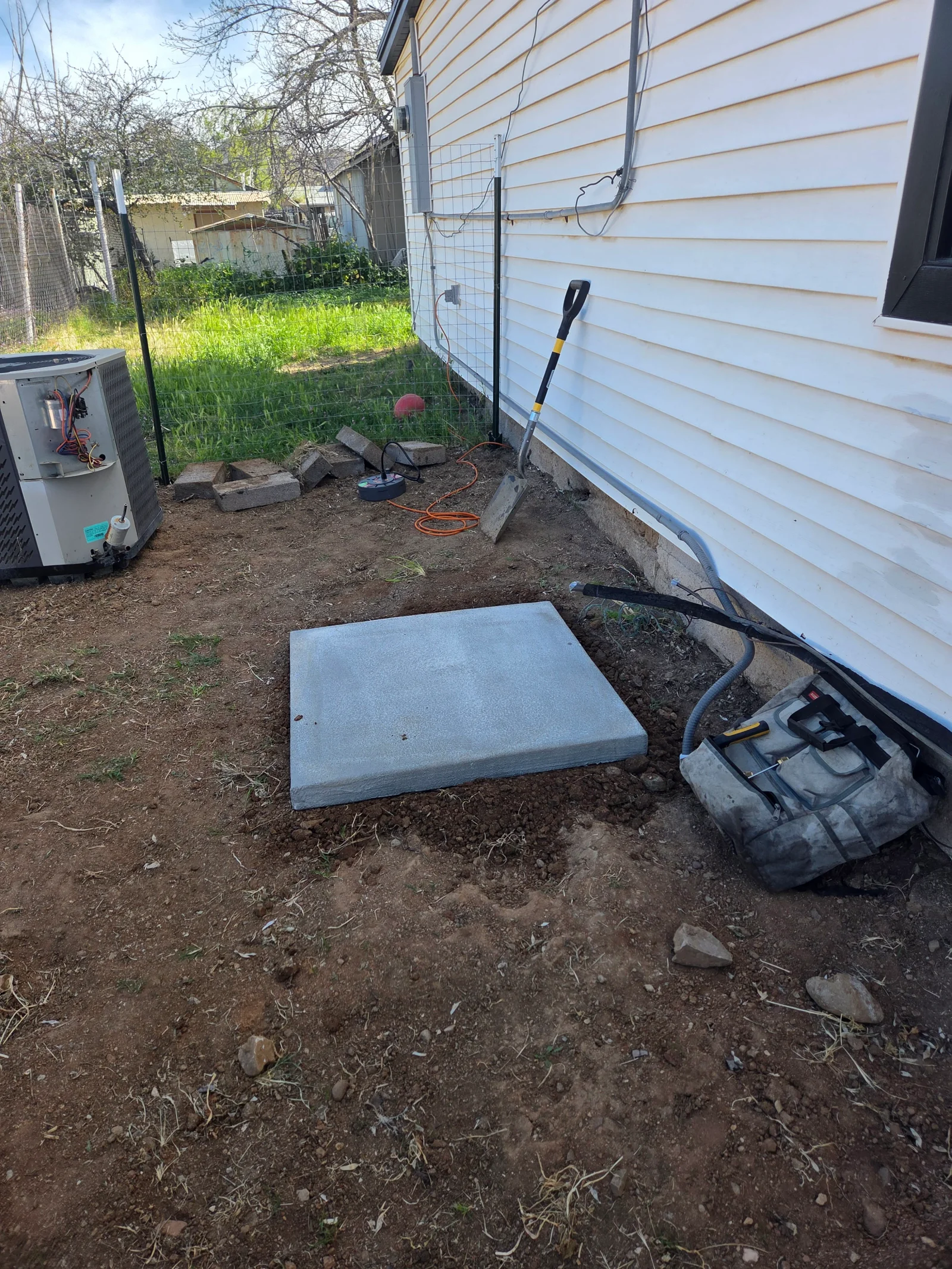 Concrete pad foundation for HVAC unit installation on prepared ground beside white vinyl siding