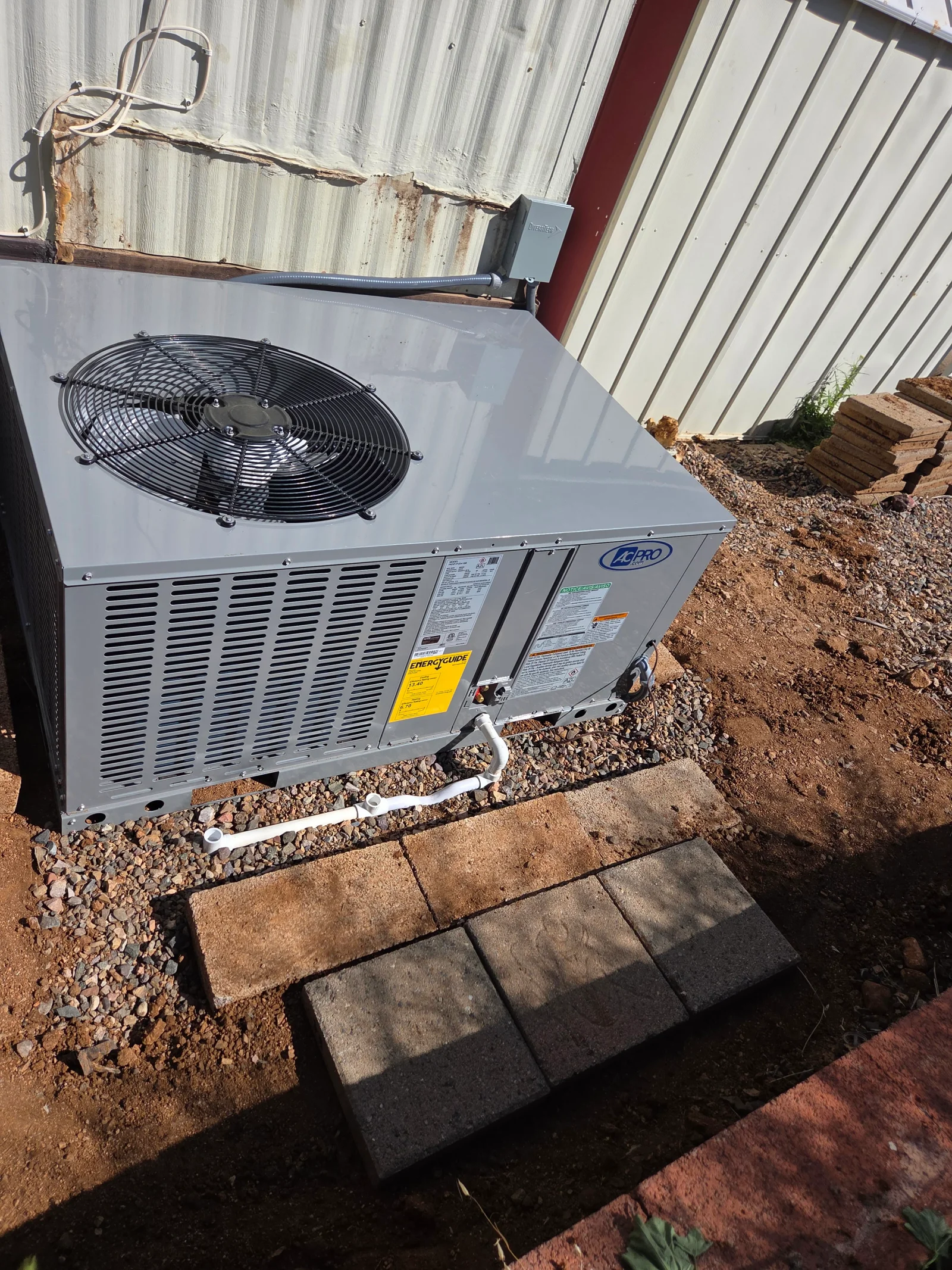 Gray packaged AC unit with black fan motor on concrete pad with gravel base and electrical connections