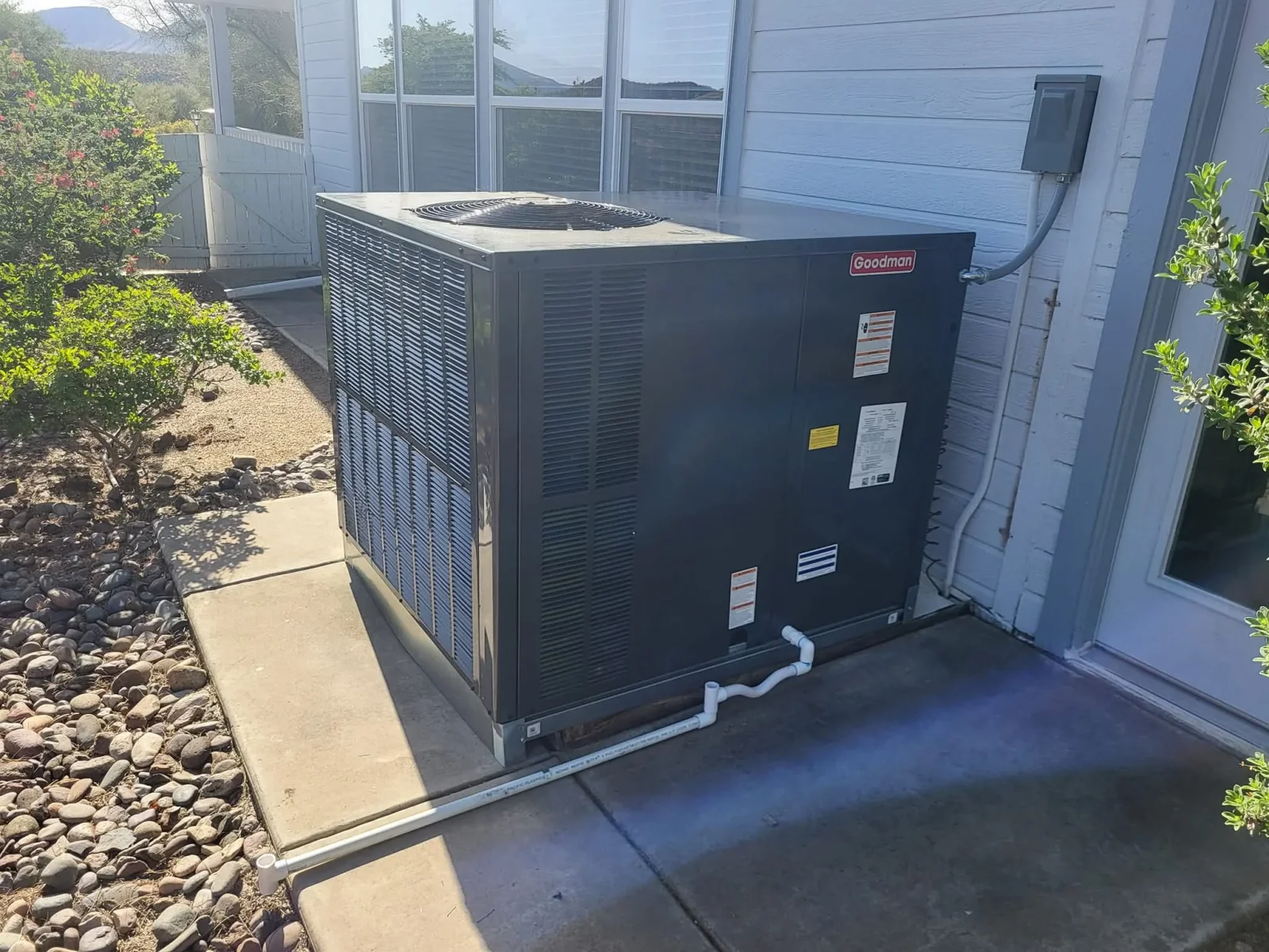 Black Goodman AC unit on concrete pad with PVC drain lines and rock landscaping