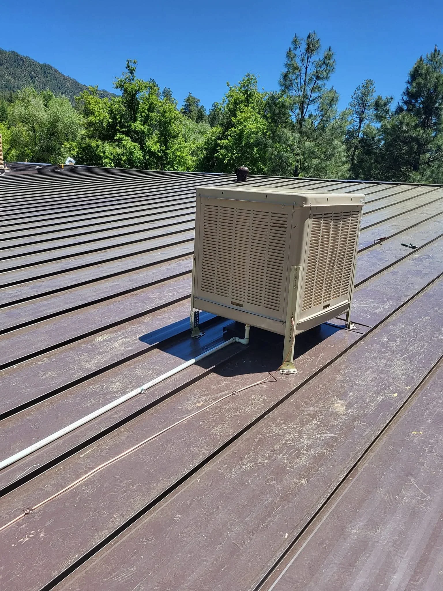 Beige evaporative cooler mounted on blue base atop standing seam metal roof with integrated ductwork